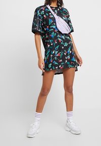 Czarna, oversize sukienka-tunika z kolorowymi kwiatowymi wzorami, krótkimi rękawami i luźnym krojem, zestawiona z białymi trampkami i paskowanymi skarpetkami.