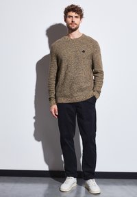 Hnědý, texturovaný pletený svetr s kulatým výstřihem, který je kombinován s tmavými kalhotami a bílými teniskami. Na hrudi se nachází malé černé logo.