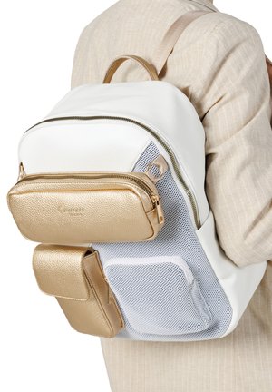 Sac à dos blanc avec poches avant dorées et poche latérale en mesh beige porté par une personne en blazer beige.