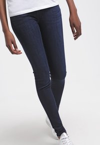 Jeans ajustados de denim azul oscuro, con una textura suave y un diseño clásico de cinco bolsillos, combinados con zapatillas blancas.