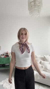 Weißes, geripptes bauchfreies Oberteil, schwarze Hose und ein rosa gemusterter Schal, der um den Hals gebunden ist. Heller Hintergrund mit Kissen und einem grünen Schrank.