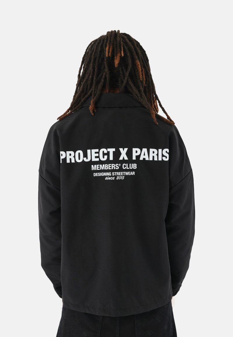 Chaqueta negra con un corte relajado. Gran texto blanco en la parte posterior que dice "PROJECT X PARIS", con detalles de marca adicionales debajo.