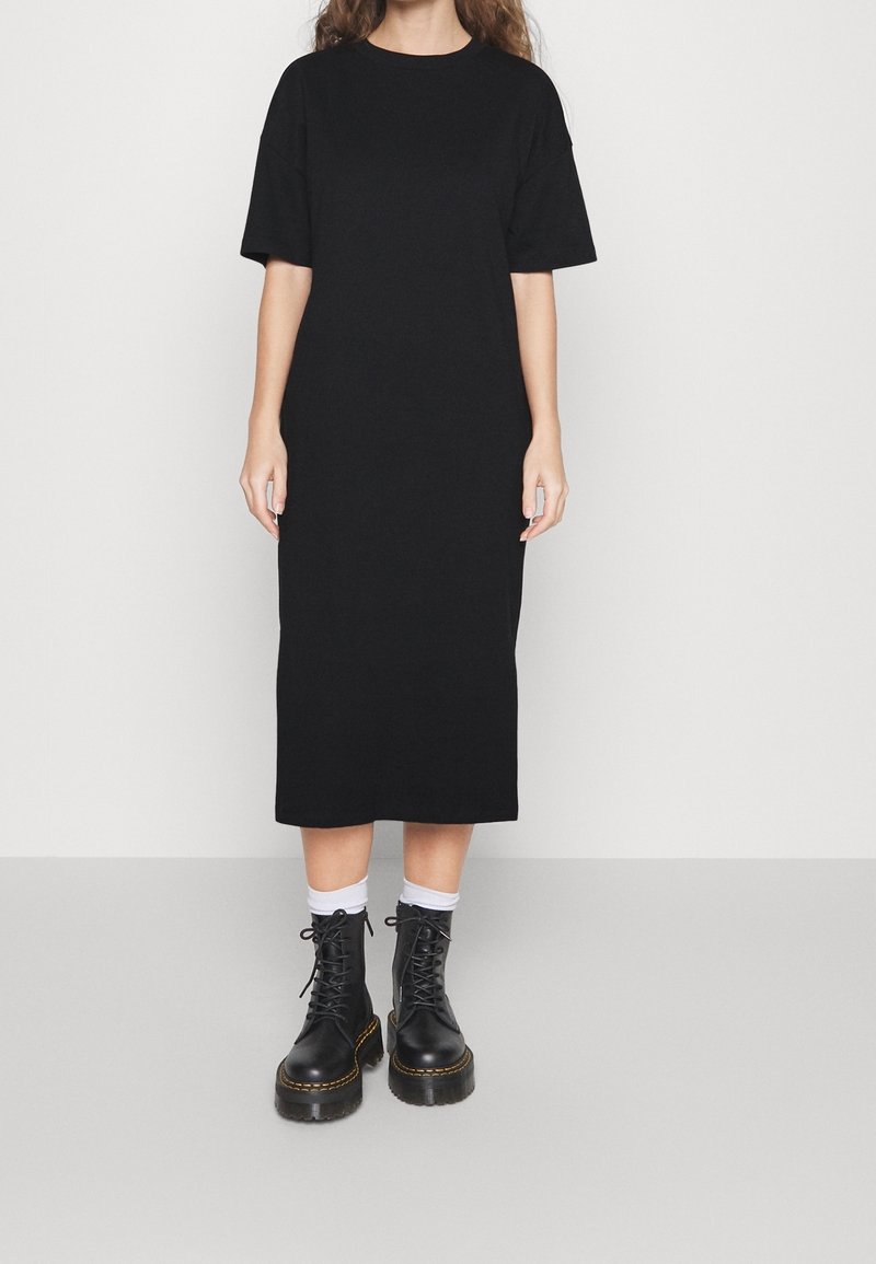 Personne portant une robe midi noire à manches courtes, des chaussettes blanches aux chevilles et des bottines noires à plateforme lacées sur un fond uni.