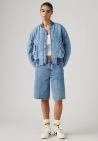 Γυναίκα που φοράει ανοιχτό μπλε τζιν μπουφάν βόμβερ και τζιν σορτς μέχρι το γόνατο με λευκά αθλητικά παπούτσια και ριγέ κάλτσες, στέκεται μπροστά σε γκρι φόντο.