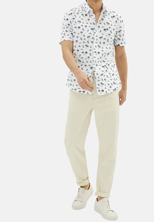 Mann trägt weißes kurzärmeliges Hemd mit Palmenmuster, beige Hose mit umgeschlagenen Bündchen an den Knöcheln und weiße Turnschuhe, steht.