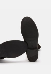 Suelas de zapatos negras con patrones de agarre texturizados, superficies lisas y un diseño minimalista. Los lados muestran un ligero desgaste y detalles de marca.