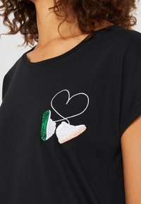 Svart t-shirt med en broderad design av två sneakers med gröna och vita accenter, sammanflätade med en vit hjärtformad linje.