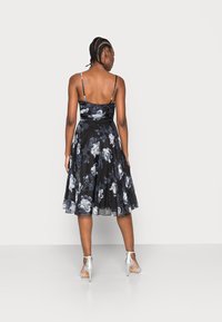 Vestido preto com padrão floral, com alças finas, até ao joelho, feito de tecido leve. Sapatos de salto prata completam o look.