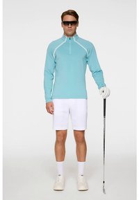 Ljusblå golftröja med lång ärm och vita kontrastsömmar, kombinerad med vita shorts och vita sneakers. Håller i en golfklubba och bär solglasögon.