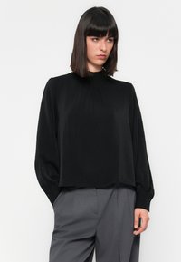Donna che indossa una blusa nera a maniche lunghe con polsini arricciati e pantaloni grigi a vita alta, in piedi di fronte a uno sfondo bianco uniforme.