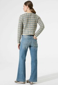 Mujer con coleta castaña vestida con un cárdigan azul de estampado, vaqueros de tiro alto azul claro y zapatos metálicos de slip-on, de pie con una mano en el bolsillo trasero.