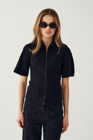 Donna con capelli lunghi indossa una maglia nera a maniche corte con zip, pantaloni neri e occhiali da sole neri stretti, in piedi contro uno sfondo semplice.