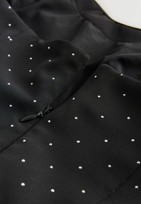 Melns audums ar maziem, vienmērīgi izvietotiem baltiem pērļu pērlēm un daļēji atvērtu melnu rāvējslēdzēju centrā.