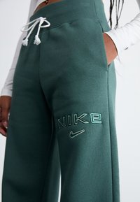 Zelené tepláky s elastickým pasem a stahovací šňůrkou. Obsahují nápis "NIKE" v texturované výšivce a malý logo Nike. Součástí jsou kapsy na bocích.