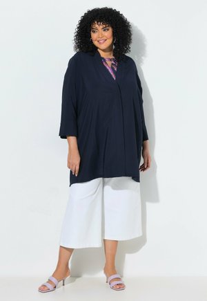 Donna con capelli ricci che indossa una maglietta larga blu navy, pantaloni bianchi a gamba larga e corti e sandali con tacco viola chiaro, in piedi contro un muro bianco.