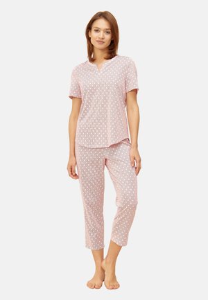 Frau steht barfuß und trägt ein kurzärmeliges, rosa Oberteil und eine kurz geschnittene Schlafanzughose mit weißen Punkten auf hellem Hintergrund.