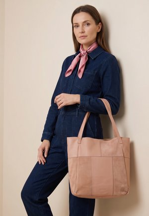 Kvinde i mørk denim-dress og lyserødt tørklæde står op ad væggen og holder en stor lyserød patchwork lædertaske.