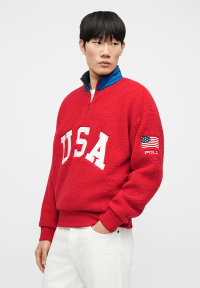 Κόκκινο φλις πουλόβερ με φερμουάρ στο ένα τέταρτο, με λευκά γράμματα "USA" και μπαλώματα με την αμερικανική σημαία στο μανίκι. Μπλε εσωτερικό γιακά.