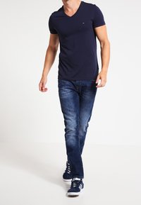 Camiseta de cuello en V azul marino, ajustada, combinada con jeans azules desgastados. Las zapatillas son de color marino con acentos blancos. El sujeto está caminando hacia adelante.