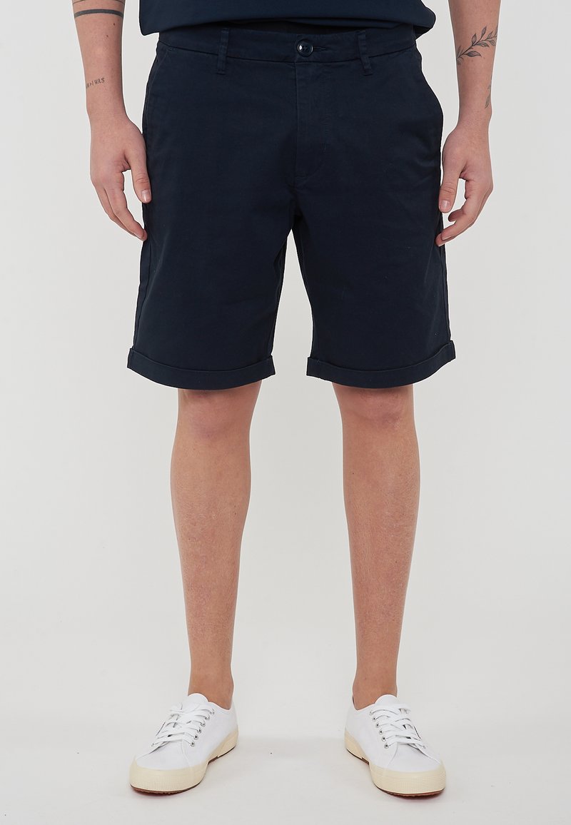 Shorts in cotone blu con orli risvoltati, tasche frontali e chiusura con un bottone. Indossati con sneakers bianche.