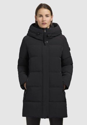 Vrouw in een zwarte gewatteerde winterjas met een hoge kraag en capuchon, met haar handen in de zakken, recht voor zich uit kijkend tegen een effen achtergrond.