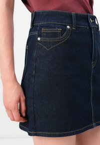 Kvinne i en mørkeblå denimskjørt med gule sømmer og frontlommer, som står mot en hvit bakgrunn.