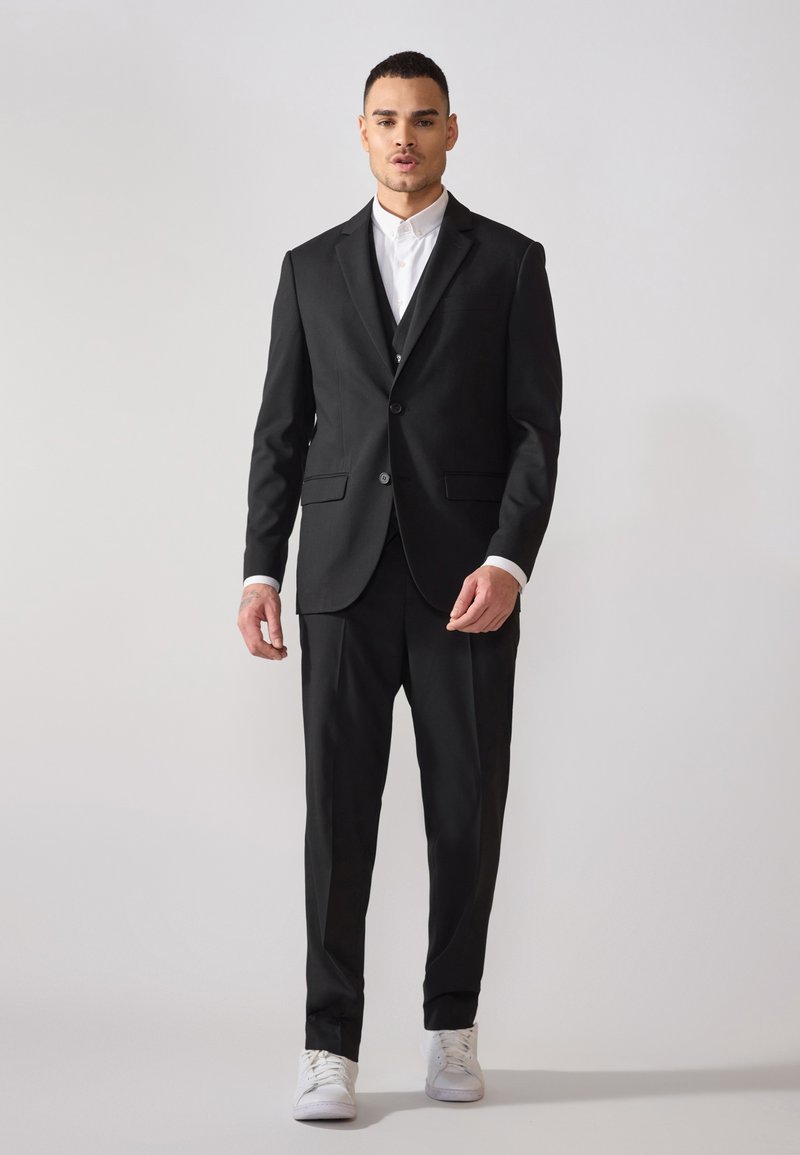 Schwarzer Anzug aus glattem Stoff mit zwei Knöpfen, schmalem Revers und slim-fit Hosen, kombiniert mit einem weißen Hemd und Sneakers.