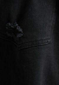 Närbild av svart denimtyg med en trasig reva nära en sydd söm, vilket framhäver textur och slitage.