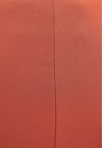 Tkanina v rezavé barvě s hladkým povrchem; má jemný lesk a vertikální šev, který probíhá středem, což vytváří štíhlý vzhled.