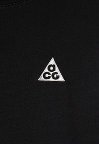 Fekete, sima textúrális anyagon egy fehér, hímzett háromszög alakú logó található, amelyben középen az "acg" felirat szerepel.