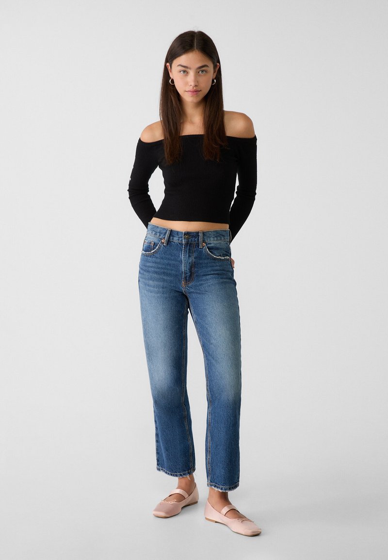 Giovane donna in piedi con le mani dietro la schiena, indossando un top lungo a maniche nere scoperte sulle spalle, jeans blu e ballerine rosa su uno sfondo semplice.