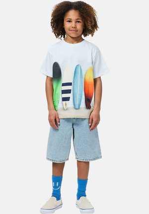 T-shirt blanc à manches courtes avec imprimés colorés de planches de surf, associé à un short en jean bleu clair et des chaussettes bleues à motifs.