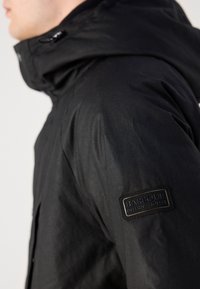 Fekete kapucnis dzseki strukturált anyagból, sima felülettel. Jellemzője egy „Barbour International” felirattal bélyegzett bőr folt az ujjon.