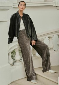 Zwarte leren cropped jas, witte top en grijze velours brede broek. Model draagt zilveren sneakers en staat op een trap.