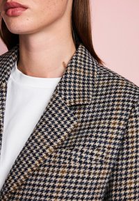 Rudo šaškinio rašto švarkas, pagamintas iš rudos, tamsiai mėlynos ir kreminės spalvų, turintis išpjautą apykaklę, tekstūruotą audinį ir uždėtas ant baltos marškinių.