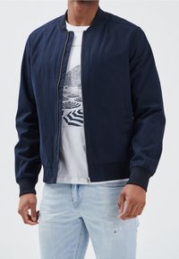 Marinblå bomberjacka i slät tyg, med dragkedja fram, ribbade ärmar och en klassisk kragsdesign, bärs över en t-shirt med tryck.