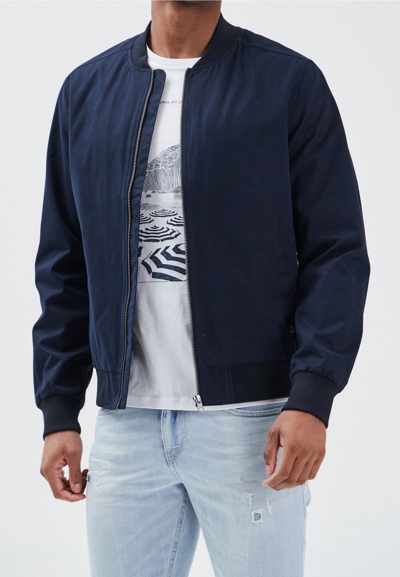 Marinblå bomberjacka i slät tyg, med dragkedja fram, ribbade ärmar och en klassisk kragsdesign, bärs över en t-shirt med tryck.