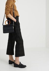 Černé krátké kalhoty s károvaným vzorem v hnědých a červených liniích, doplněné huňatým hořčicovým svetrem a černými slip-on botami. Strukturovaná černá kabelka má kruhové oranžové ucho.