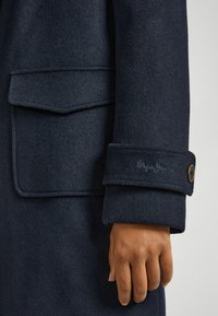 Primo piano di una mano che tiene un cappotto blu scuro con un bottone e un logo ricamato sul polsino, mostrando la texture e i dettagli del tessuto.