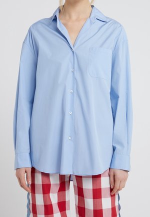 Persoon draagt een losse lichtblauwe blouse met borstzak en rood-witte geruite broek.