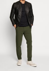 Giacca in pelle nera con girocollo, abbinata a pantaloni tapered verde scuro e sneakers bianche. Texture liscia e design minimalista.