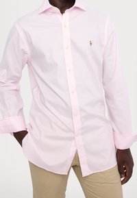 Světlé růžové košile s knoflíky a vertikálním pruhovaným vzorem, zastrčené do khaki kalhot, s dlouhými rukávy a malým logem na hrudi.