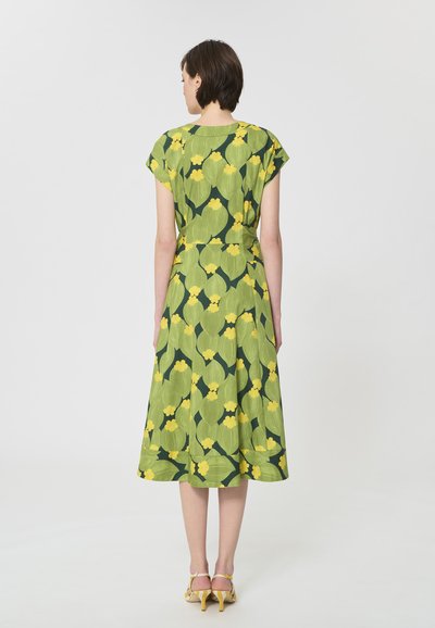 Femme debout de dos, portant une robe verte avec de grands motifs floraux jaunes et des sandales à talons hauts jaunes.