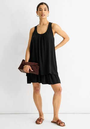 Abito nero senza maniche con tessuto fluido e design a strati. Il modello tiene una borsa a tracolla intrecciata marrone e indossa sandali marroni. Silhouette semplice e casual.