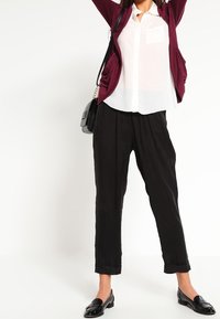 Burgundský cardigan přes bílou košili s knoflíky, černé zúžené kalhoty a černé mokasíny. Tento outfit se vyznačuje jemnými texturami a jednoduchými liniemi.
