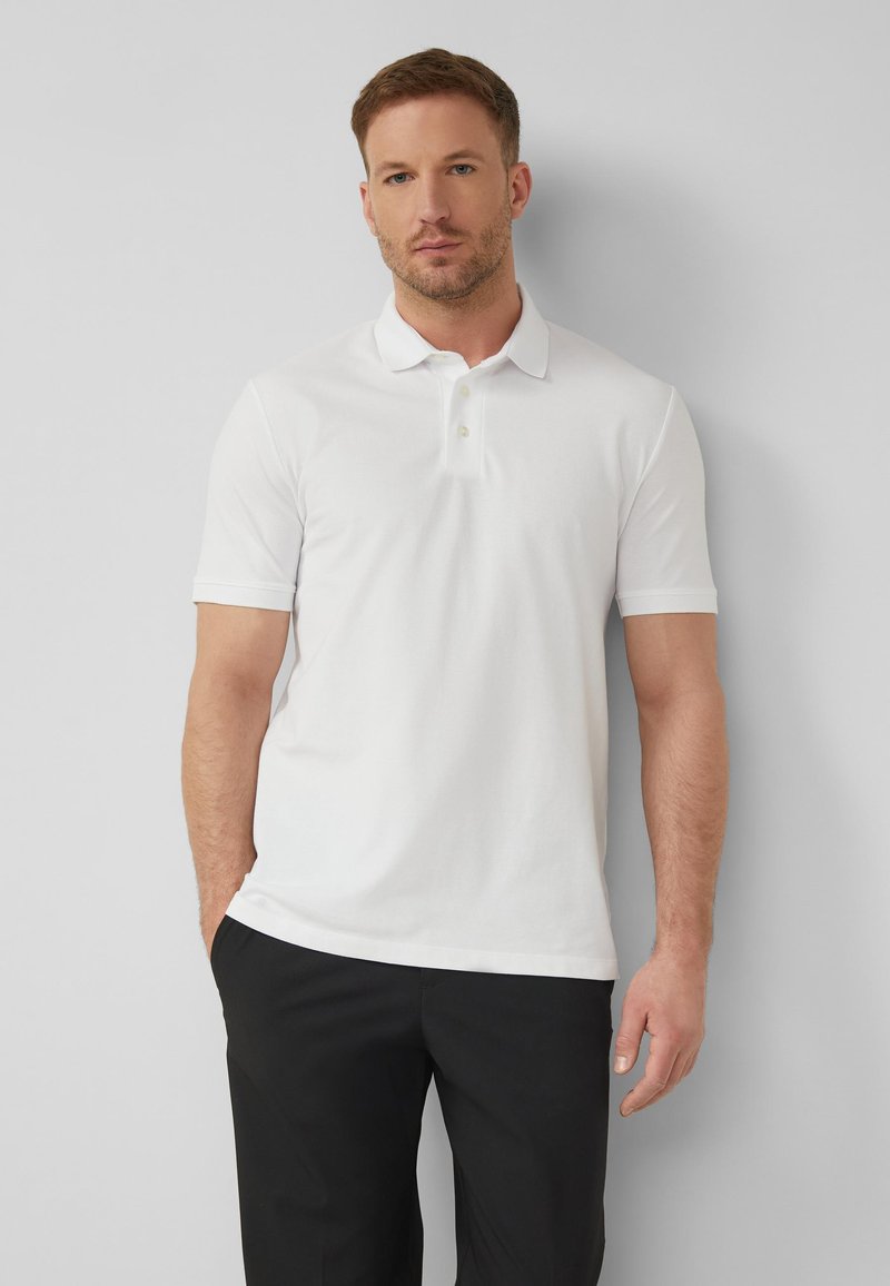 Homme portant un polo blanc à manches courtes et un pantalon noir, debout contre un mur gris clair avec une main dans la poche.