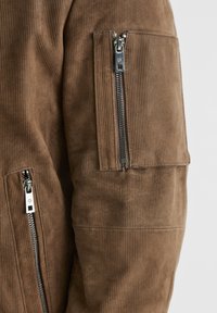 Braune Cordjacke mit silbernen Reißverschlüssen, strukturiertem Stoff und Akzenttaschen. Sichtbare Nähte und gerippte Gestaltung im gesamten Kleidungsstück.