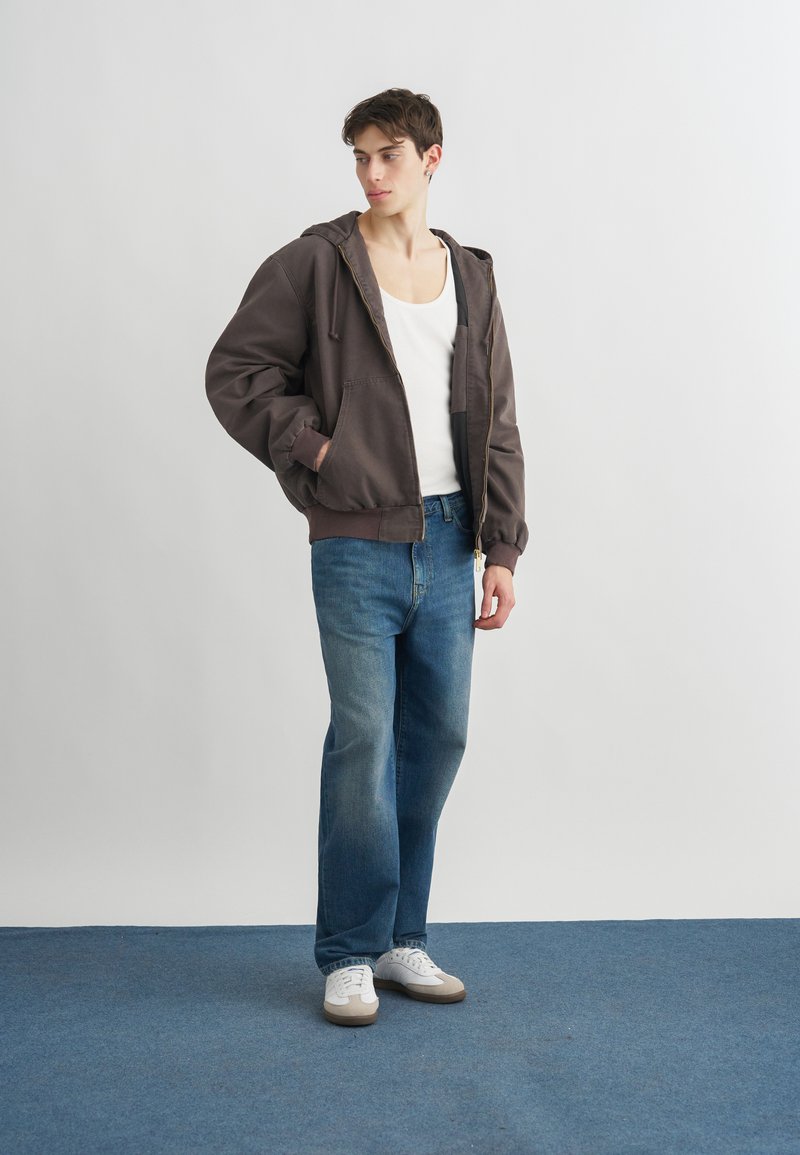 Giovane in piedi al chiuso che indossa una giacca con cappuccio marrone scuro, canottiera bianca, jeans blu e sneakers bianche, che guarda verso sinistra.