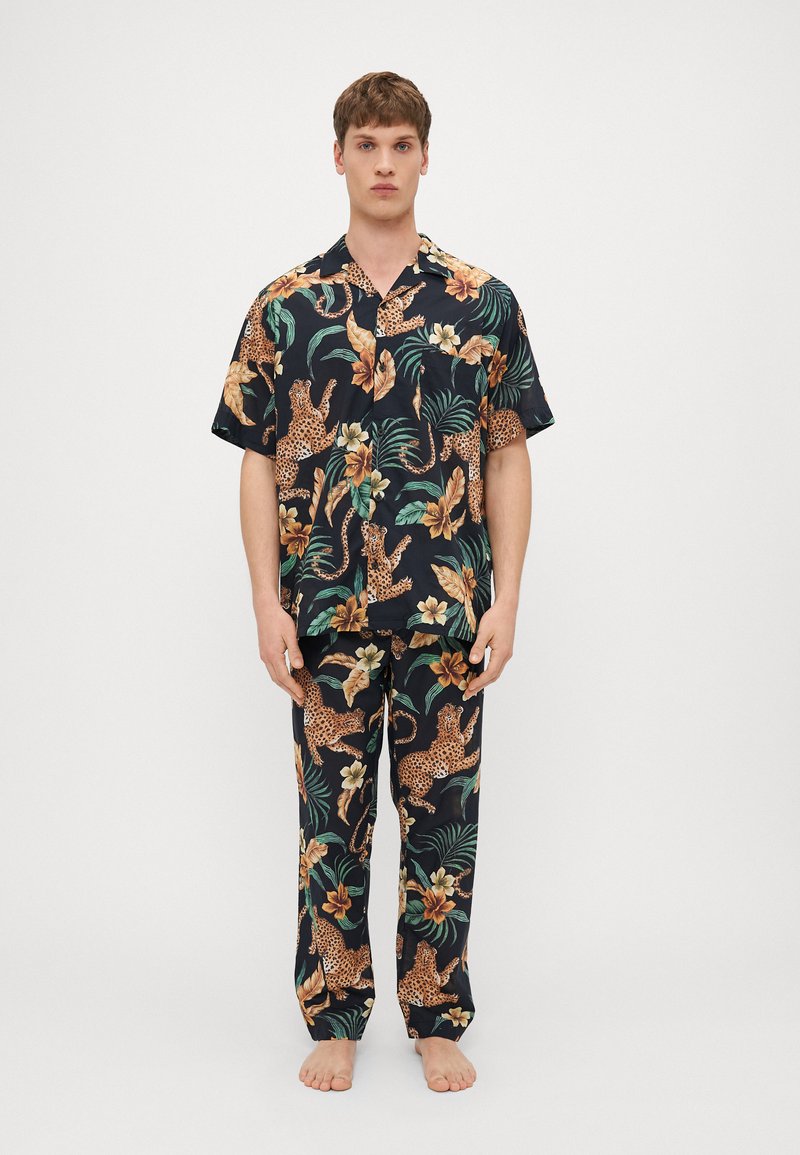 Set de pijamale cu maneca scurta, de culoare neagra, cu un imprimeu vibrant de junglă, ce include lei, frunze tropicale și flori în tonuri de portocaliu și verde.