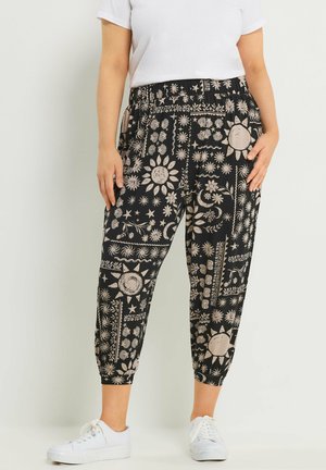 Frau trägt schwarze, gekürzte Hose mit beigen Sonnen-, Stern- und Blumenmustern, kombiniert mit weißen Turnschuhen und einem weißen Kurzarmhemd.